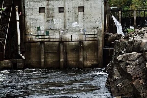 A concrete dam with graffiti, a turbulent river, and a rocky outcrop.