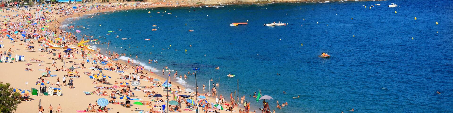 A crowded beach with clear blue water, a small island with buildings, and a clear sky.