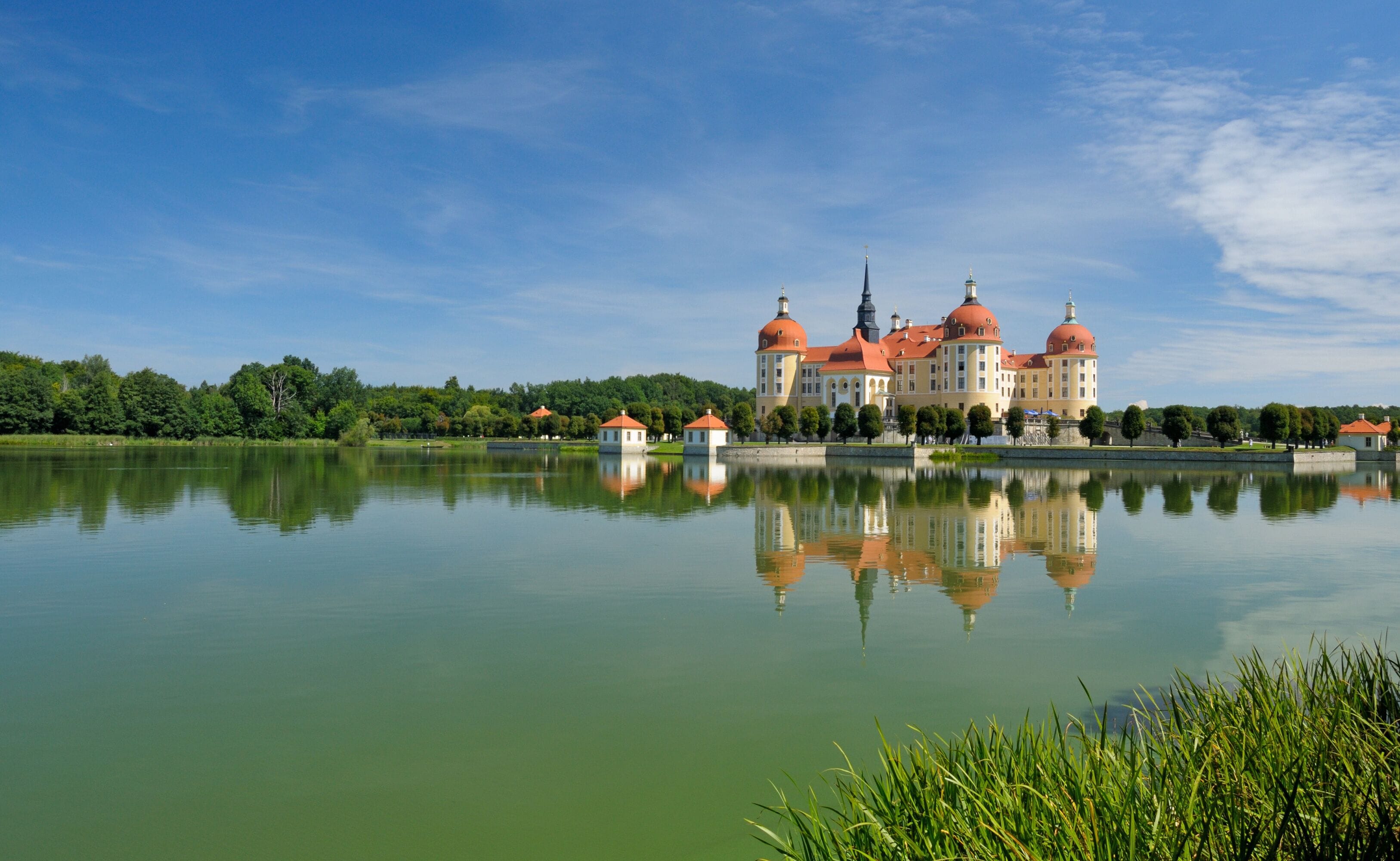 Un gran palacio con cúpulas rojas a orillas de un lago tranquilo, rodeado de árboles y un cielo despejado.