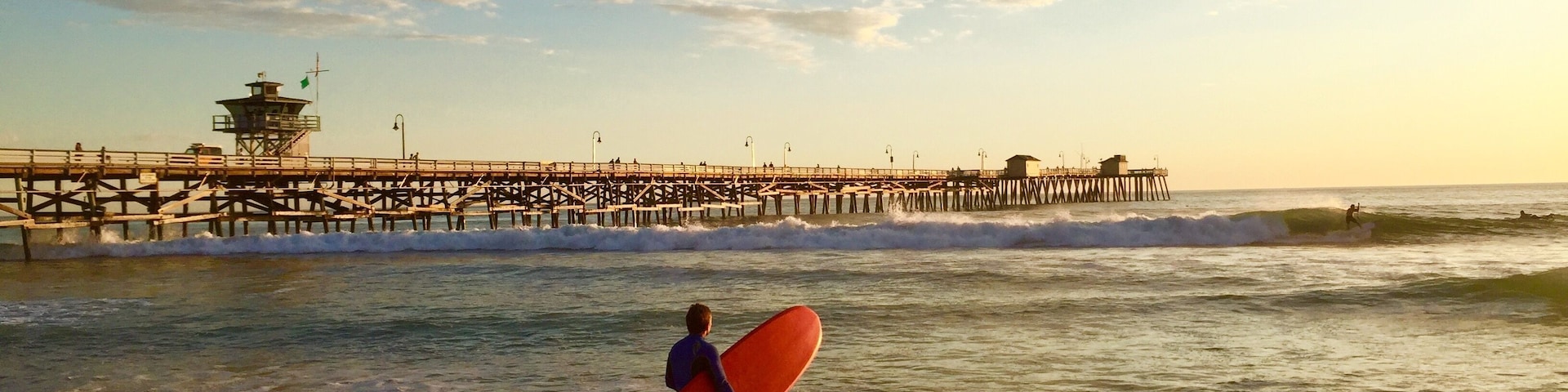 Uma praia com um cais se estendendo para o oceano, ondas quebrando na costa e uma pessoa carregando uma prancha de surf.