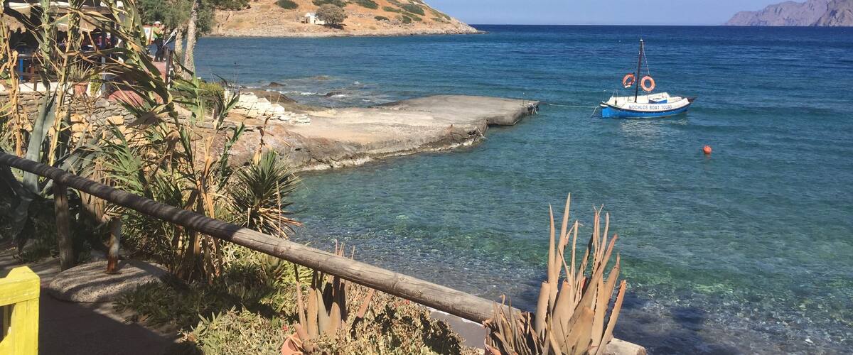 A coastal scene with a clear blue sea, a small boat named 'MOCHLOS REAL HOURS', and a rocky shoreline with vegetation.
