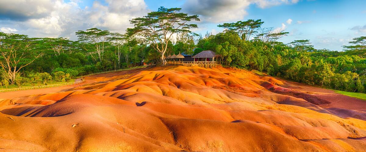 A landscape with undulating orange sand dunes, a wooden structure, and a backdrop of dense green foliage.