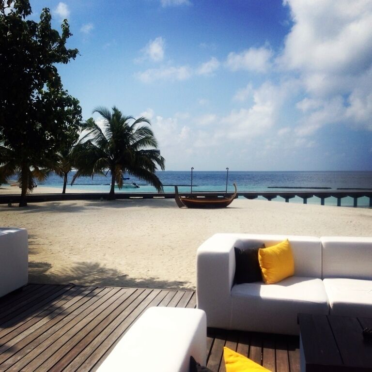 A beach with a wooden deck, a white sofa with yellow pillows, palm trees, and a boat on the water.