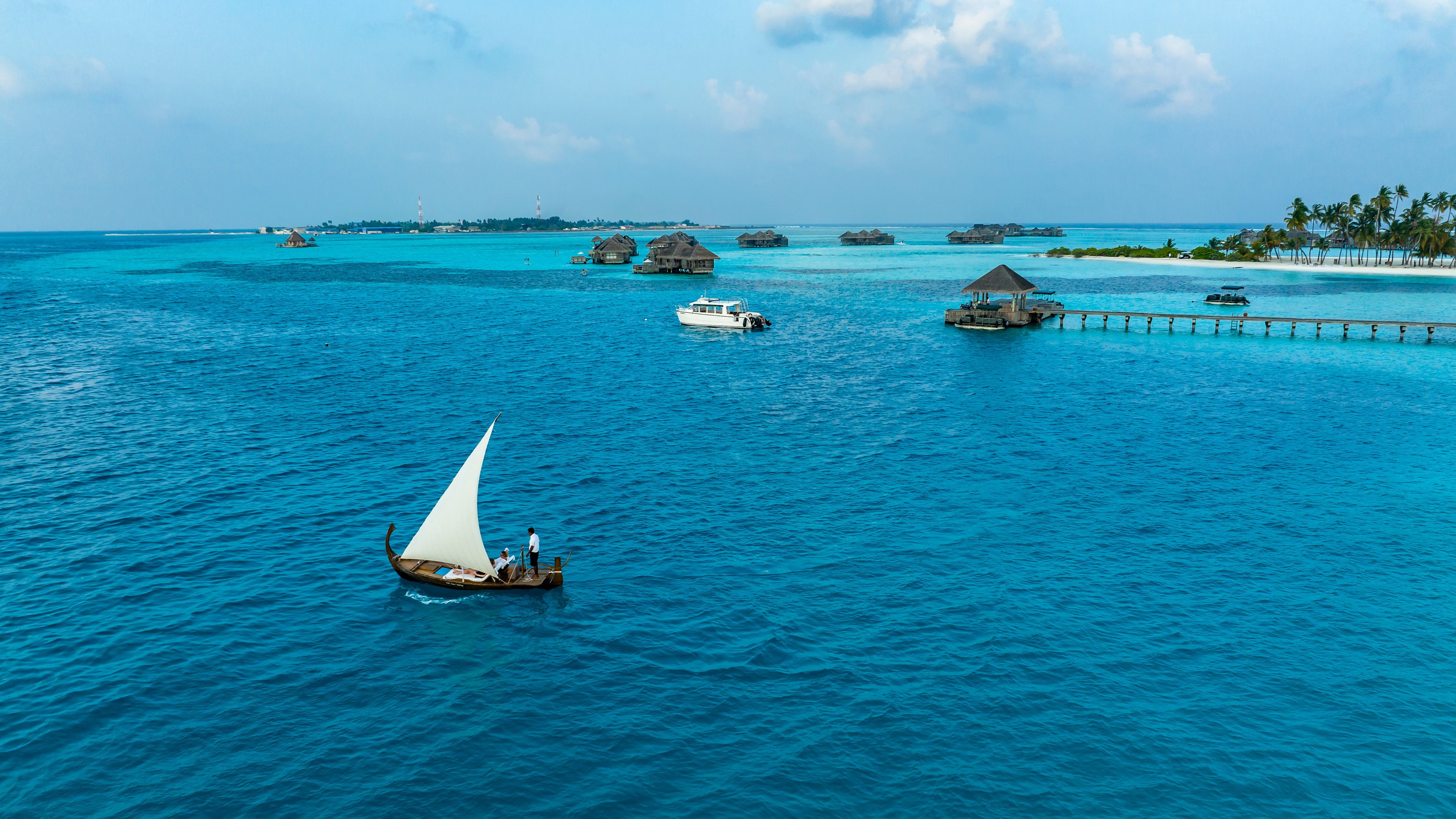 A sailboat on turquoise waters with overwater bungalows and a pier extending into the sea.