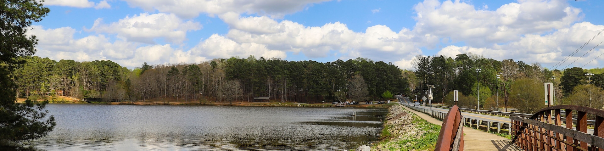 a metal and wooden rust colored bridge over a lake surrounded by lake water, green grass and lush green trees with powerful clouds and blue sky at Murphey Candler Park in Atlanta Georgia USA
