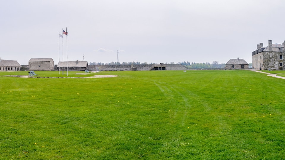 A historic fort with stone buildings, a grassy field, and a flagpole with flags.