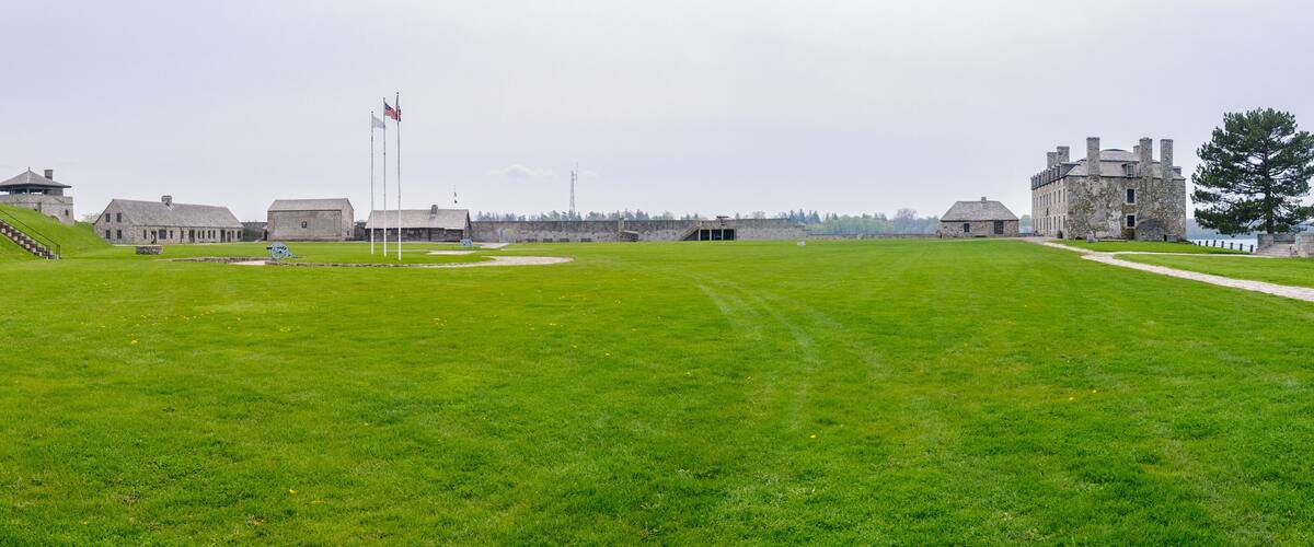 A historic fort with stone buildings, a grassy field, and a flagpole with flags.