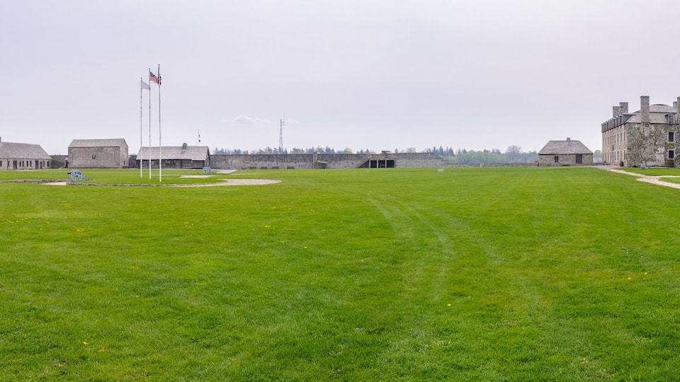 A historic fort with stone buildings, a grassy field, and a flagpole with flags.