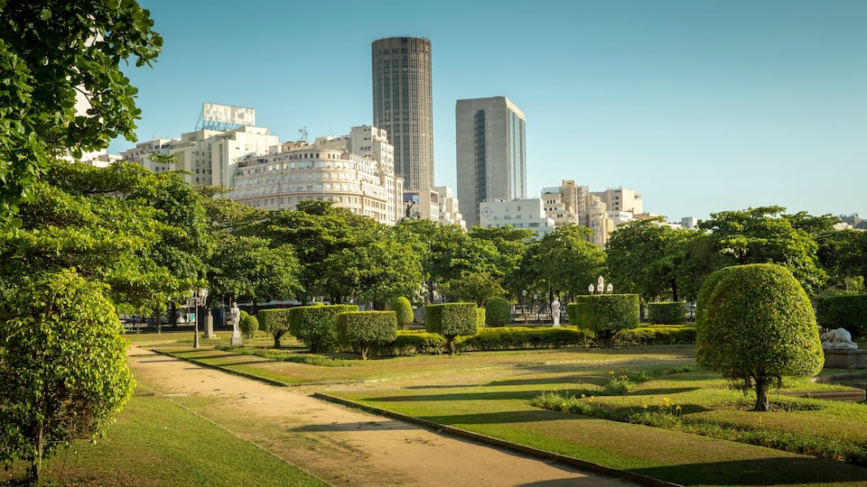 Um parque com gramados bem cuidados, sebes e uma estátua, com edifícios da cidade ao fundo.