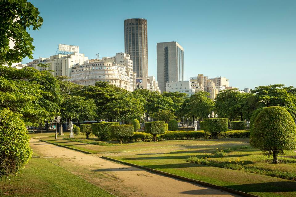 Um parque com gramados bem cuidados, sebes e uma estátua, com edifícios da cidade ao fundo.