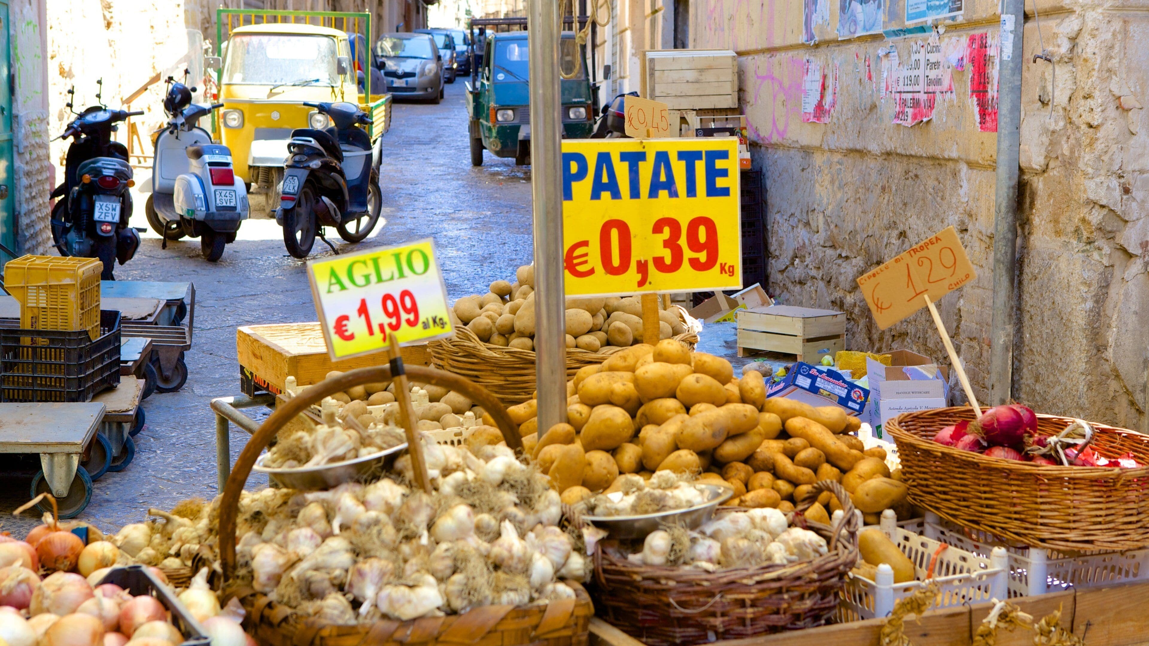 A street market with various vegetables like potatoes and onions, and signs indicating prices for potatoes and garlic.