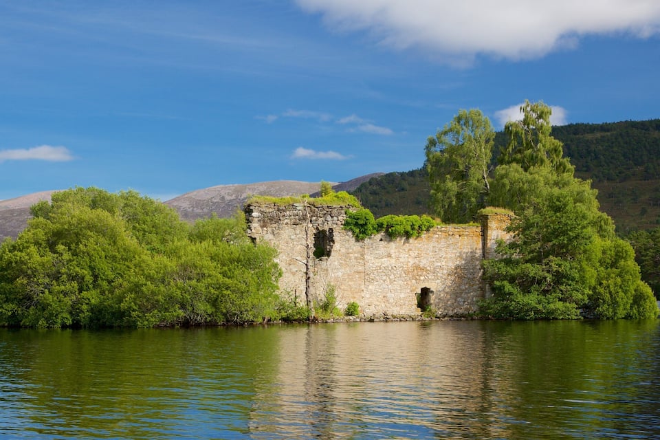 A stone ruin surrounded by lush greenery and a calm lake, with mountains in the background.