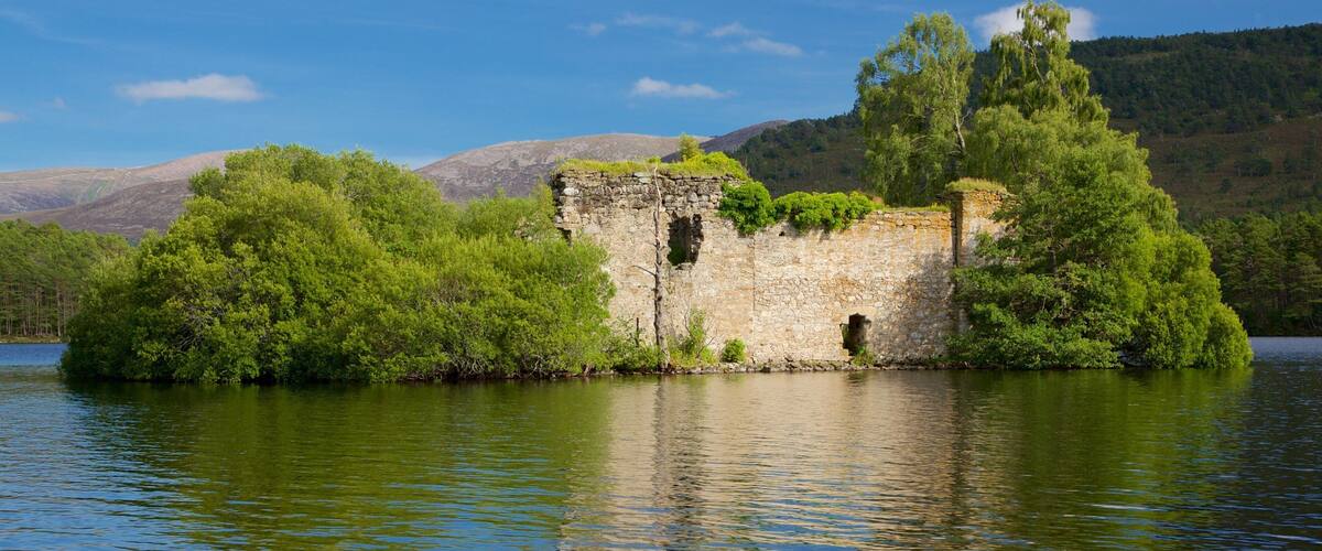 A stone ruin surrounded by lush greenery and a calm lake, with mountains in the background.