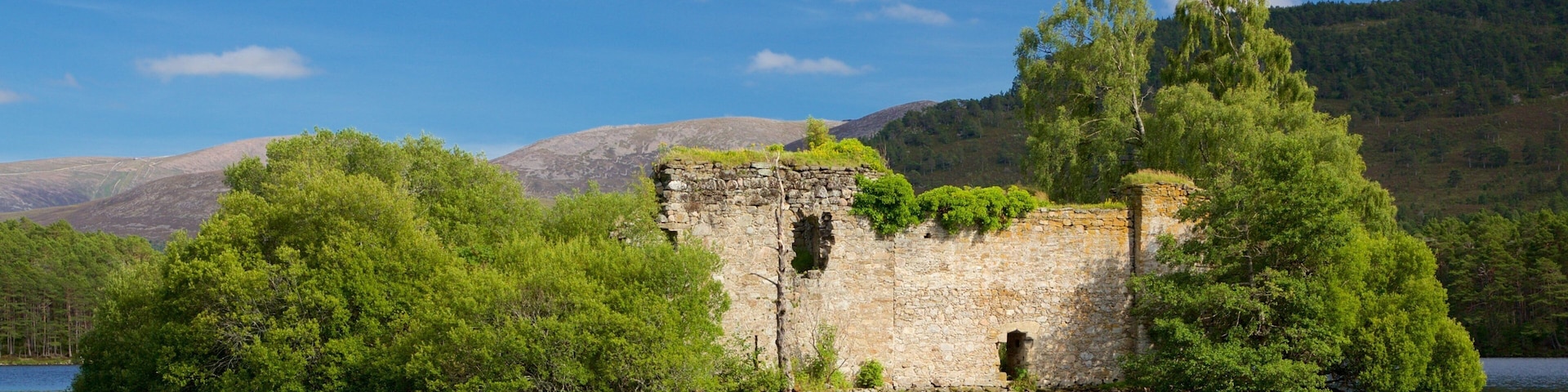 A stone ruin surrounded by lush greenery and a calm lake, with mountains in the background.