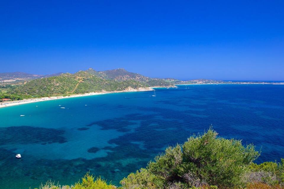 Paisaje costero con aguas turquesas cristalinas, una playa arenosa y montañas al fondo.
