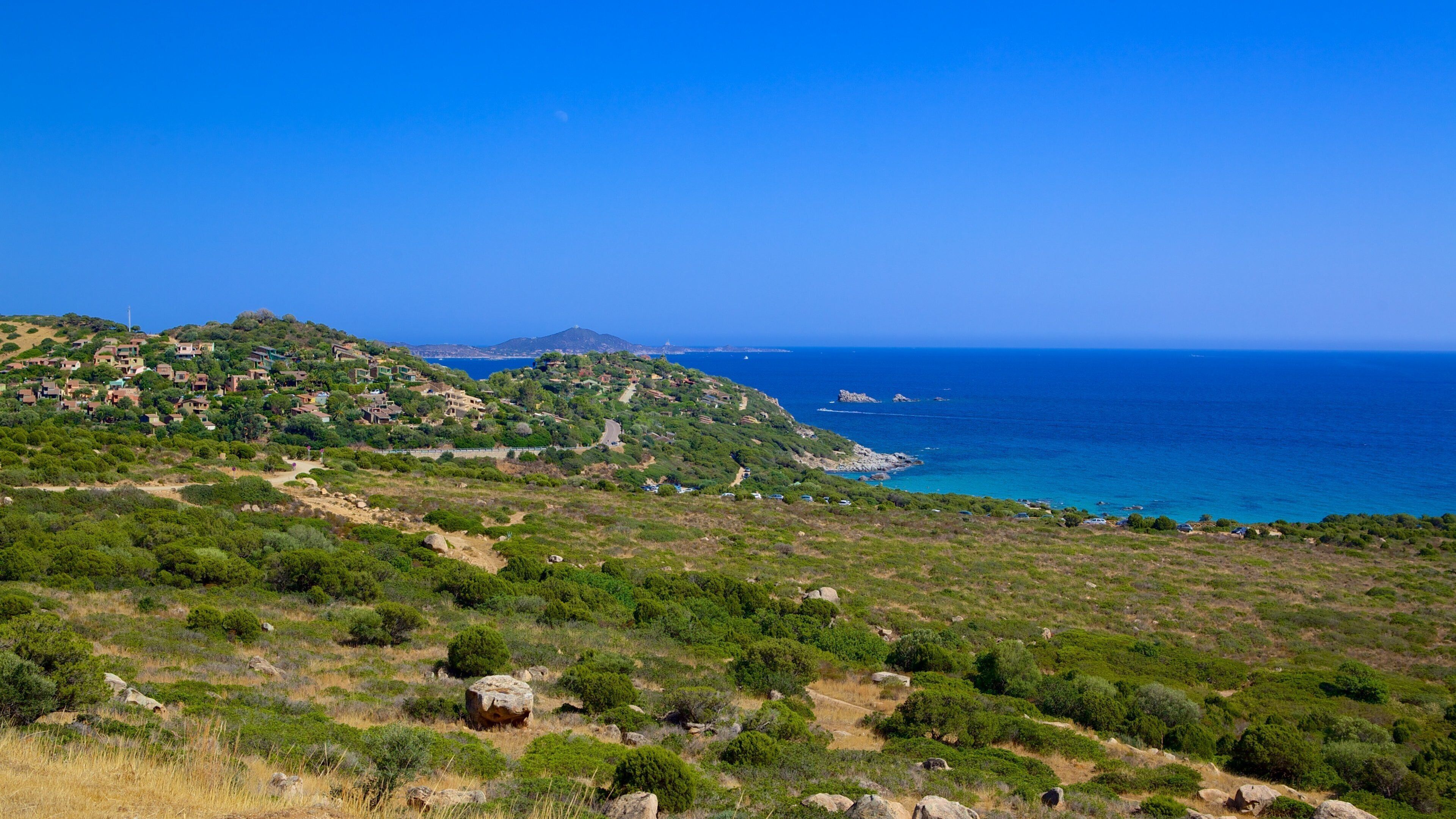 A coastal landscape with a clear blue sky, green hills, and a winding road leading to the sea.
