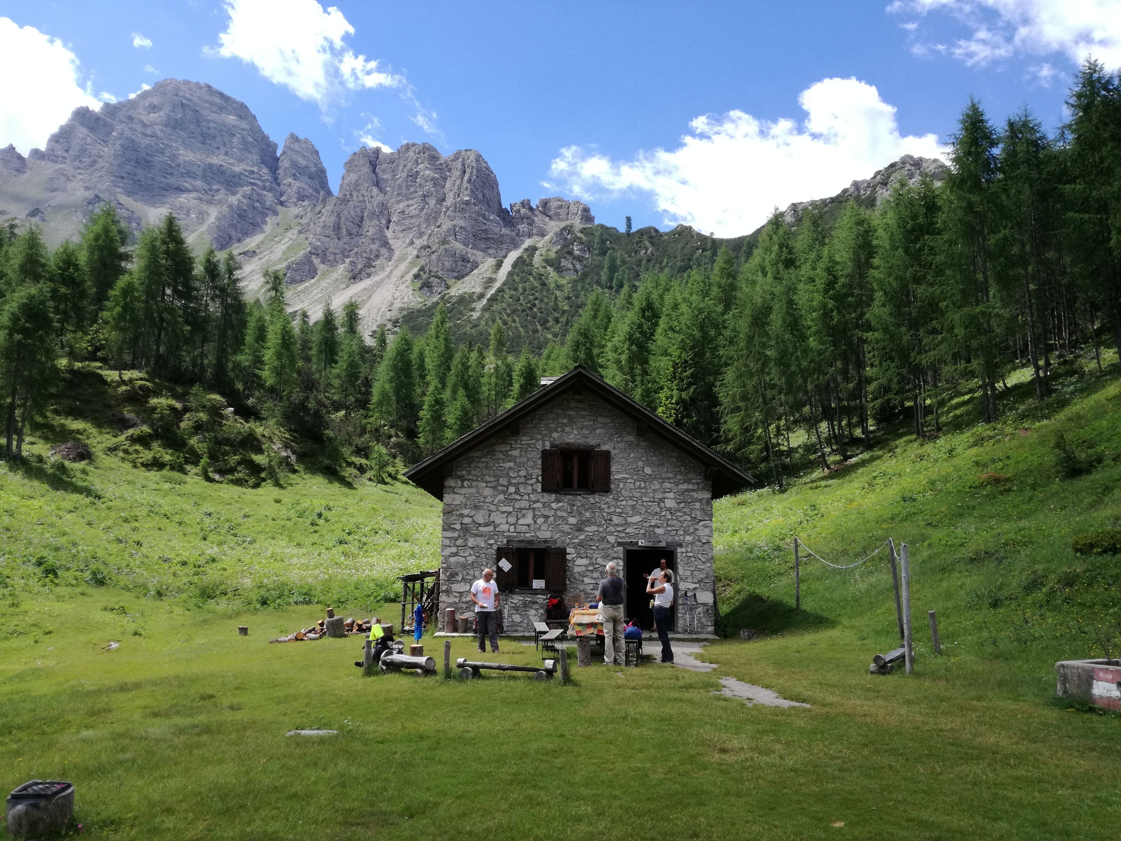 A stone cabin with a wooden door, surrounded by lush green grass and trees, with mountains in the background.
