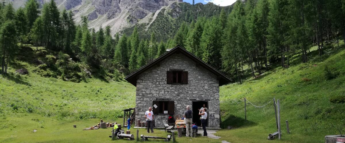 A stone cabin with a wooden door, surrounded by lush green grass and trees, with mountains in the background.
