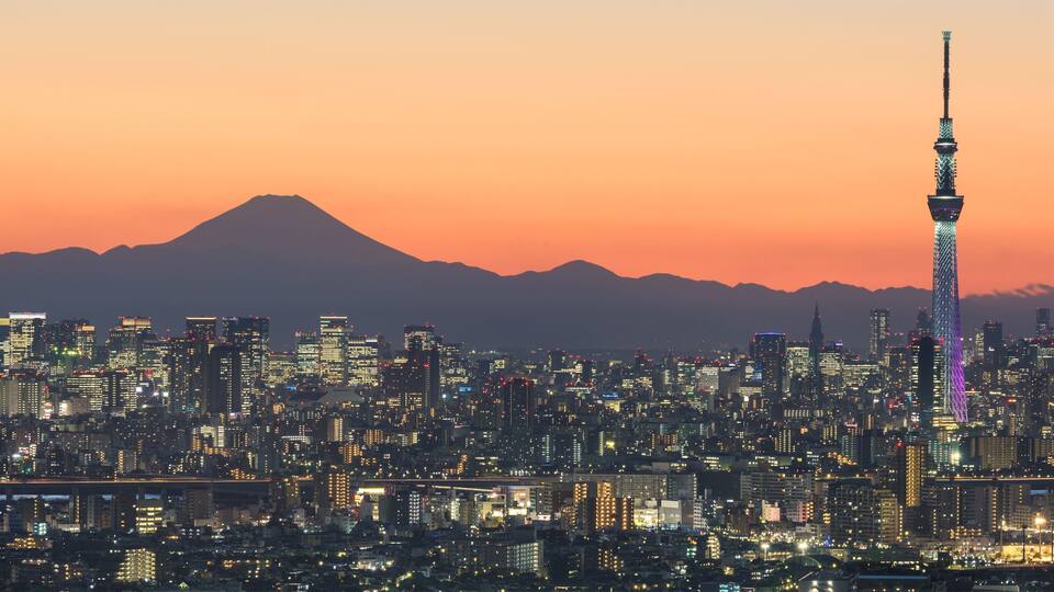 Paisagem urbana ao entardecer, com uma torre imponente e uma montanha ao fundo.