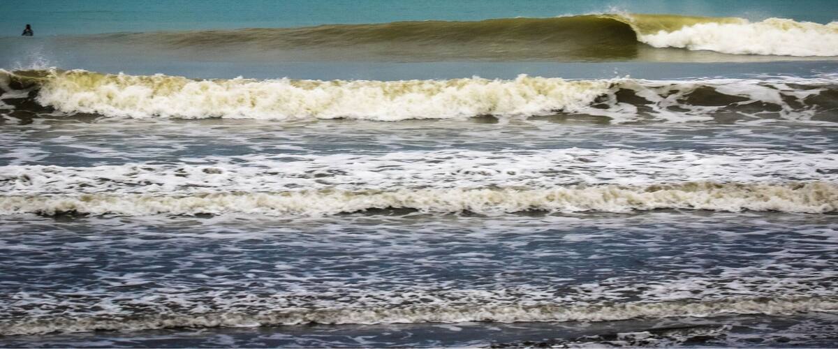 A beach with waves crashing onto the shore and a person surfing in the distance.