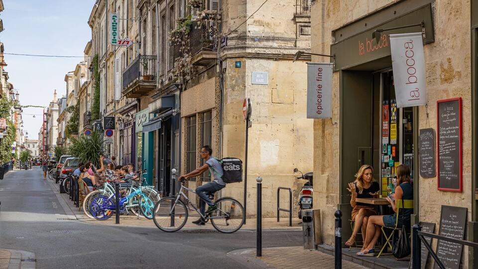 Une rue bordée de vélos garés, un homme à vélo et une femme assise à la terrasse d’un café.