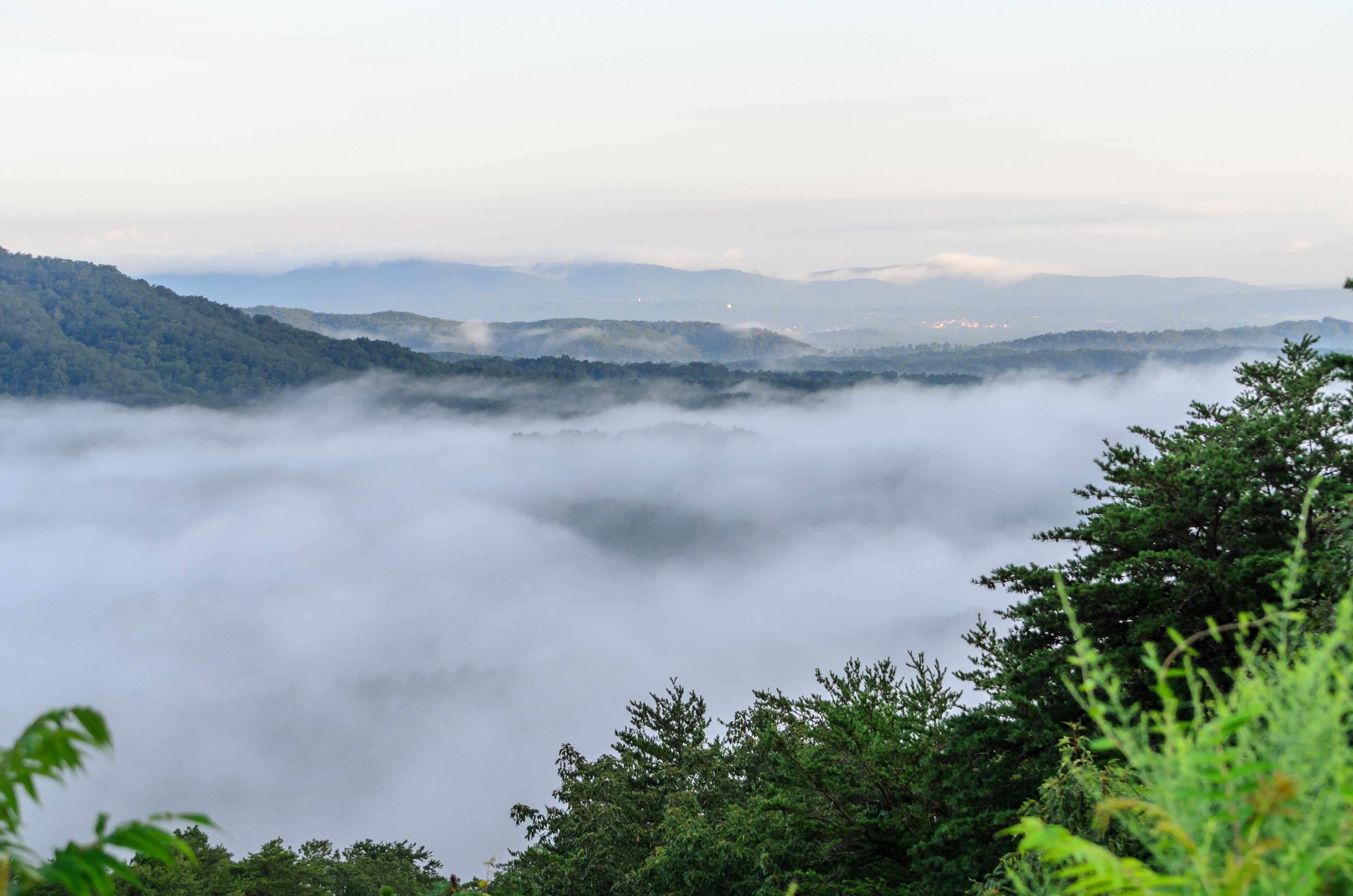 A valley with a blanket of clouds, forested hills, and distant mountains.