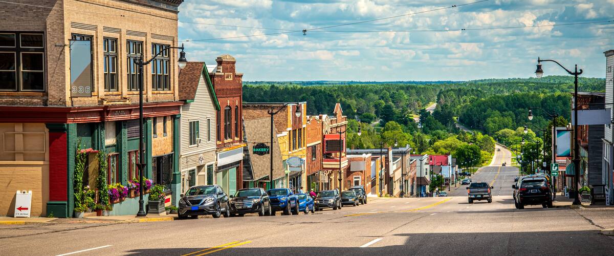 A street lined with historic buildings, parked cars, and a clear view of a forested hill in the distance.