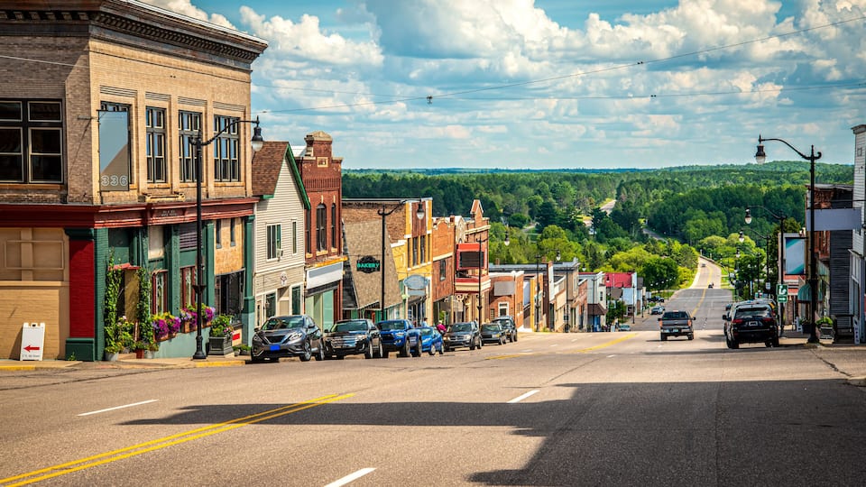 A street lined with historic buildings, parked cars, and a clear view of a forested hill in the distance.