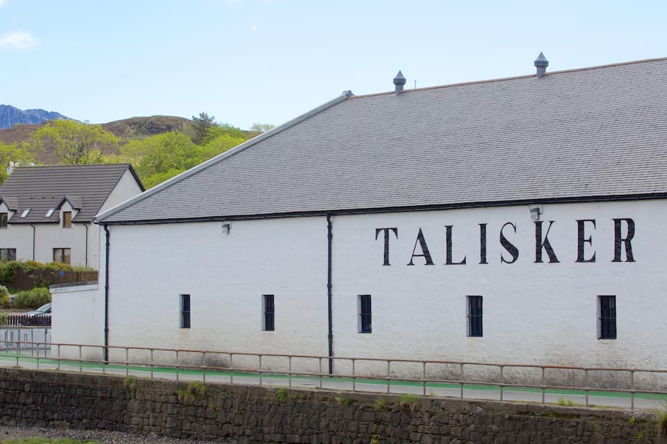 A white building with the name "TALISKER" on its side, set against a backdrop of mountains and greenery.