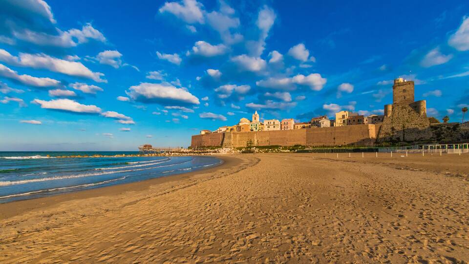 A beach with a historic castle and fortifications, a clear sky, and the ocean.