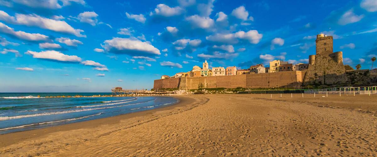 A beach with a historic castle and fortifications, a clear sky, and the ocean.