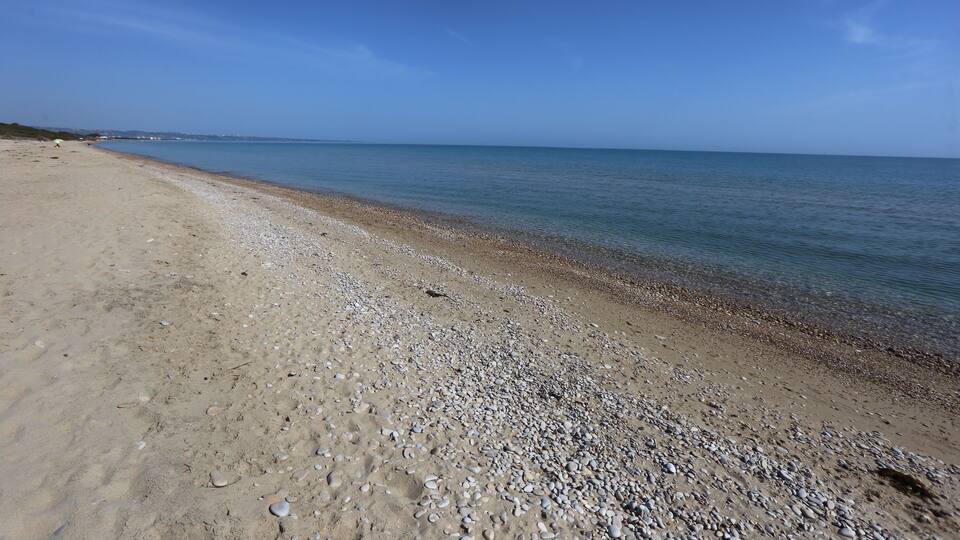 A sandy beach with pebbles and calm water under a clear blue sky.
