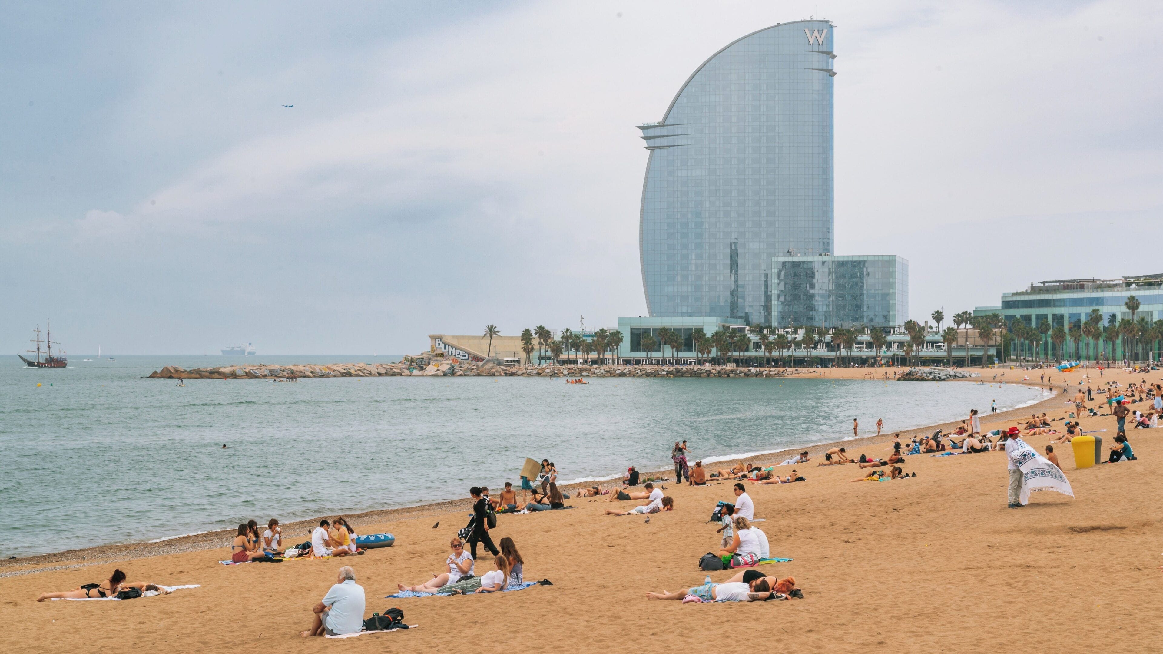 Visitors enjoy a sunny day at Barceloneta Beach in Barcelona, Catalonia, with people relaxing by the sea and the iconic W Barcelona hotel in the background
