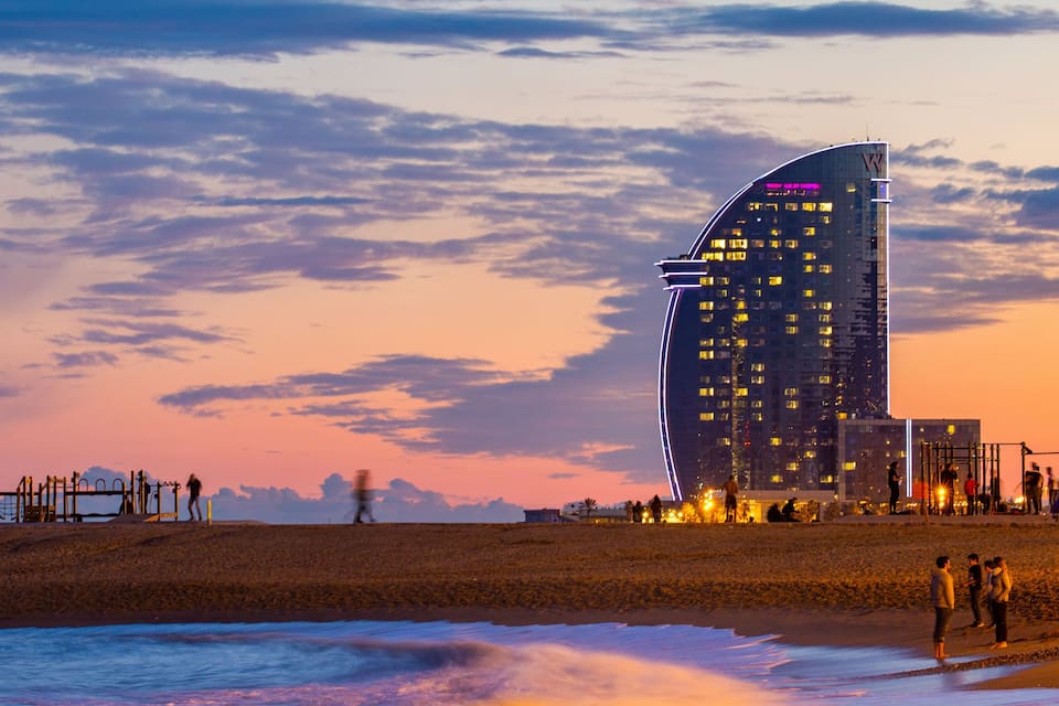 Un bâtiment moderne à l’architecture arrondie, une plage animée et un ciel au coucher du soleil.