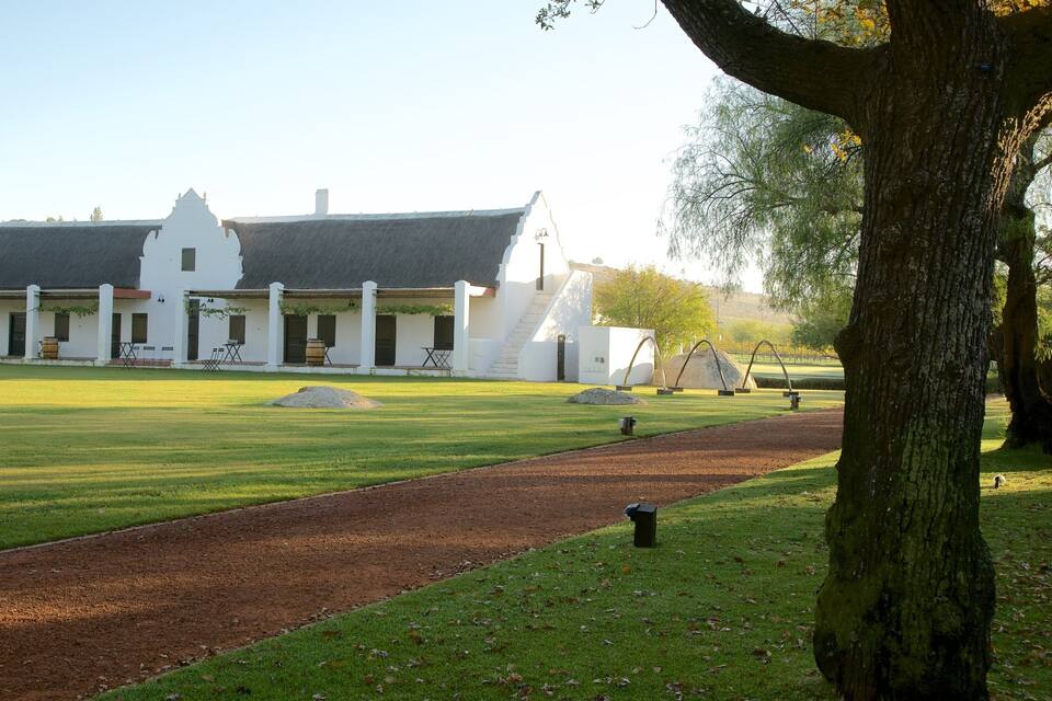 A white colonial-style building with a thatched roof, surrounded by a well-maintained lawn and a dirt path.