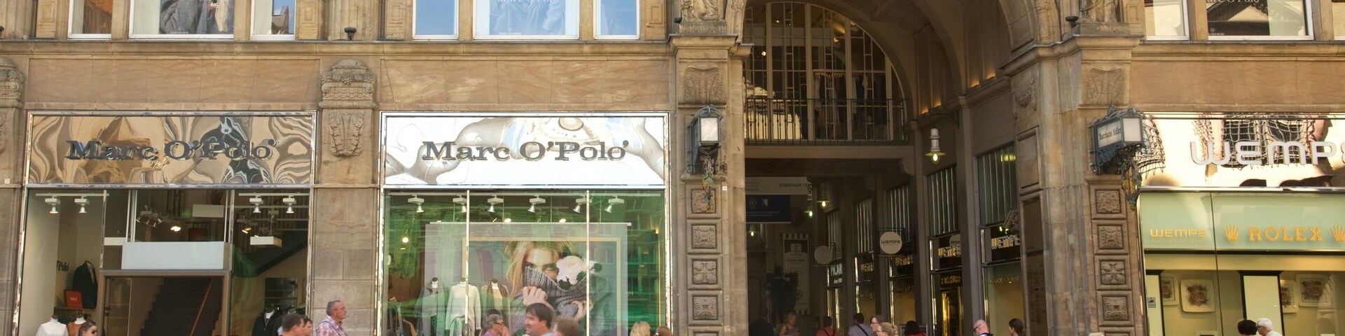 Una calle concurrida con comercios, incluyendo Marc O'Polo y Rolex.