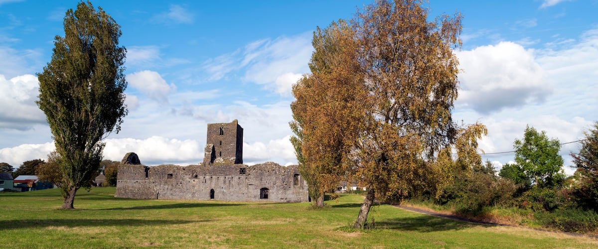 A historic stone building with a tower, surrounded by grassy fields and autumn-colored trees.