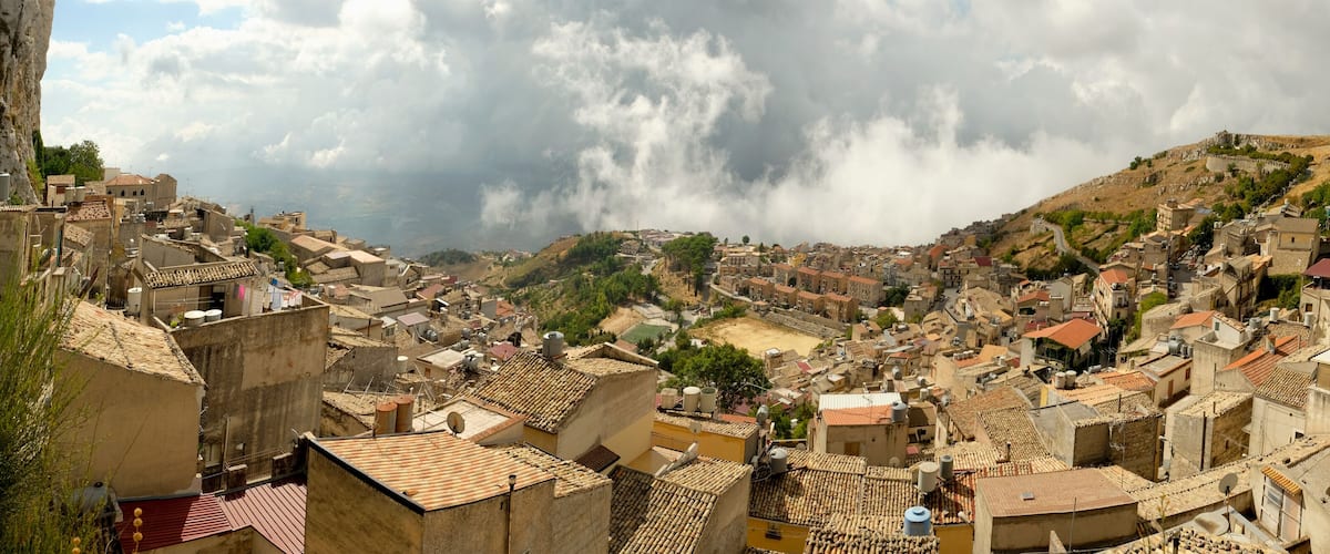 A densely packed hillside town with terracotta-roofed buildings, a prominent rocky cliff to the left, and a cloudy sky.