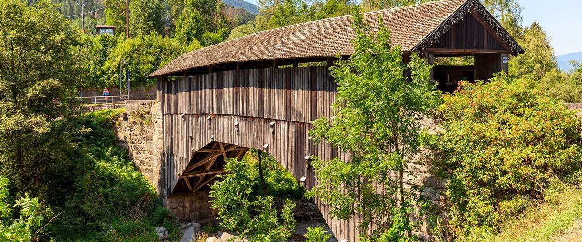 A wooden bridge with a metal arch over a stream, surrounded by lush greenery and mountains in the background.