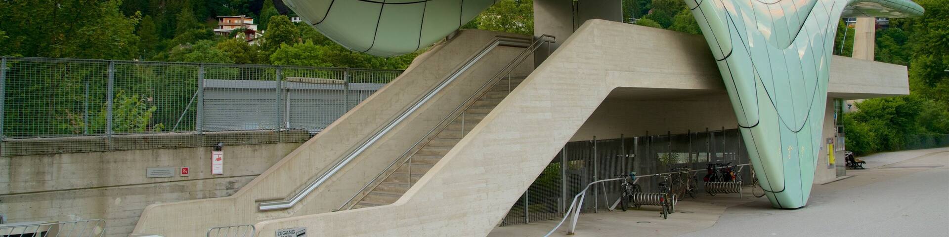 A modern architectural structure with a curved roof and stairs leading up to it, set against a mountainous backdrop.