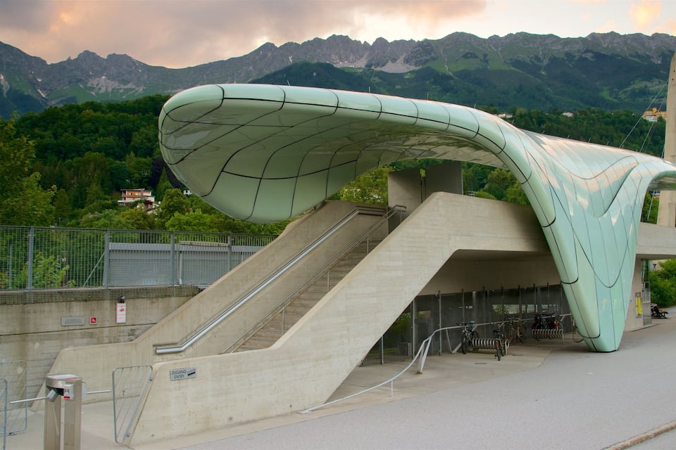 A modern architectural structure with a curved roof and stairs leading up to it, set against a mountainous backdrop.