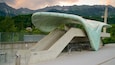 A modern architectural structure with a curved roof and stairs leading up to it, set against a mountainous backdrop.