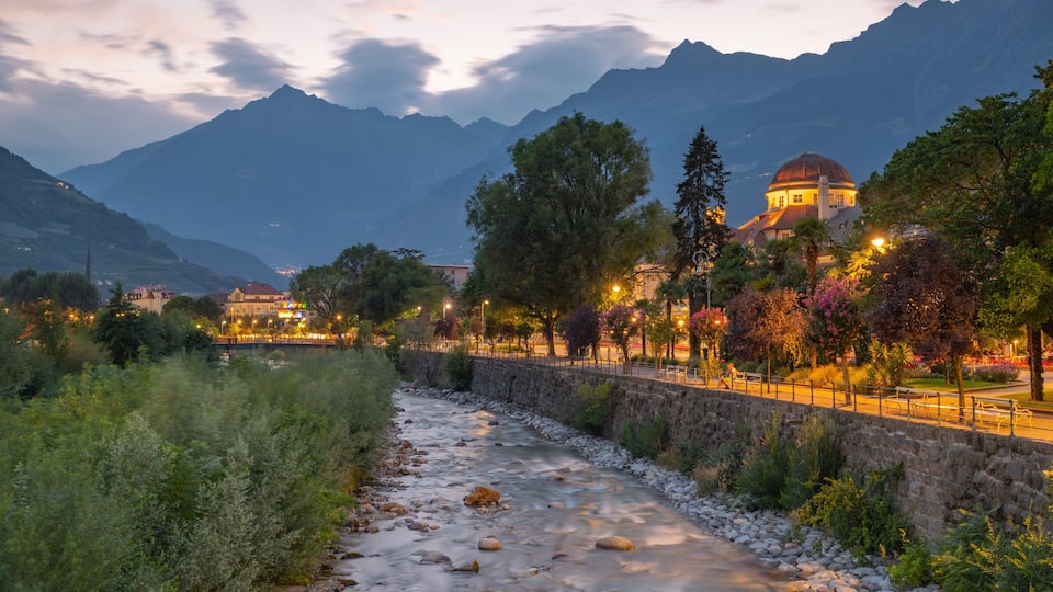 A river flowing through a town with mountains in the background and a domed building illuminated at dusk.