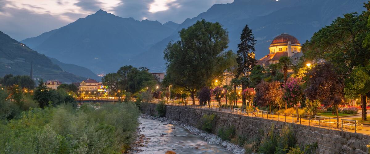 A river flowing through a town with mountains in the background and a domed building illuminated at dusk.