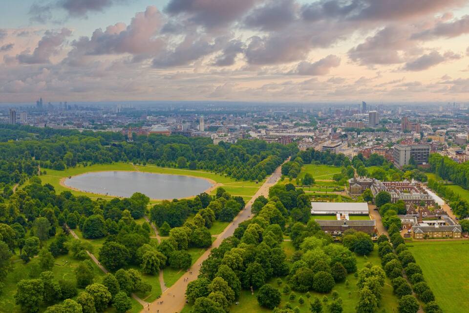 Un parque con un lago, rodeado de árboles verdes y un paisaje urbano al fondo.