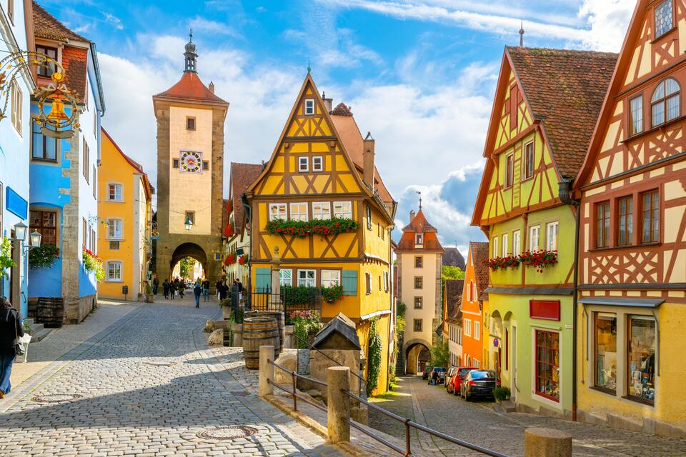 A cobblestone street lined with colorful half-timbered buildings, a tower with a clock, and an archway in the distance.