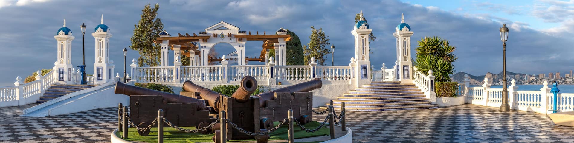 A historical monument with cannons, a checkered pavement, and a white fence with blue domes.