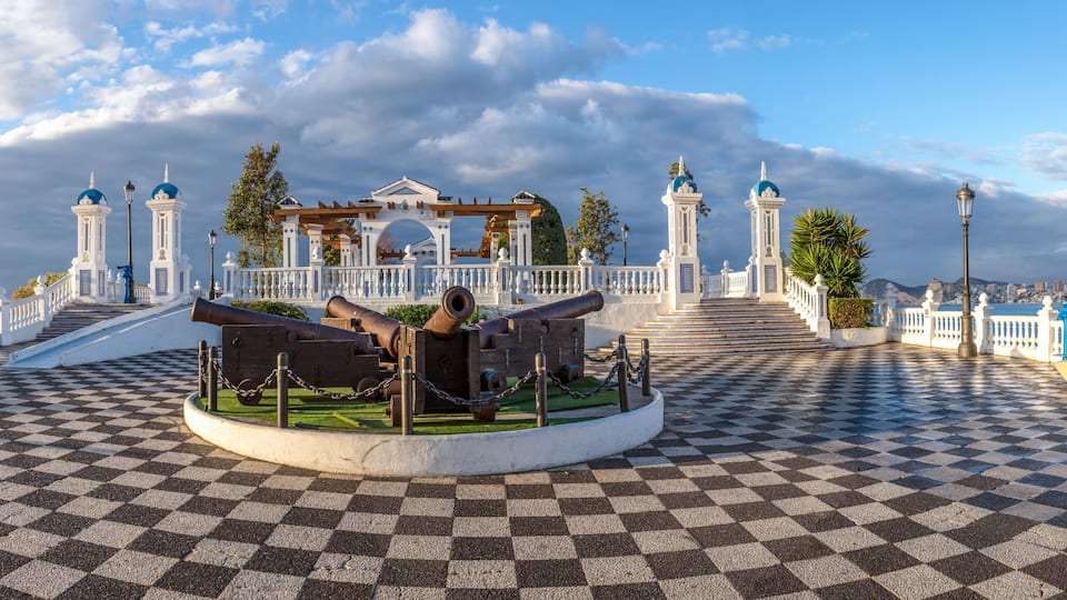 A historical monument with cannons, a checkered pavement, and a white fence with blue domes.