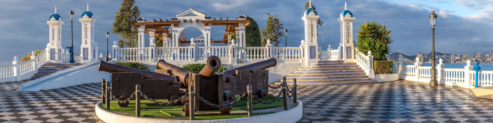 Un monumento histórico con cañones, adoquinado a cuadros y una cerca blanca con cúpulas azules.