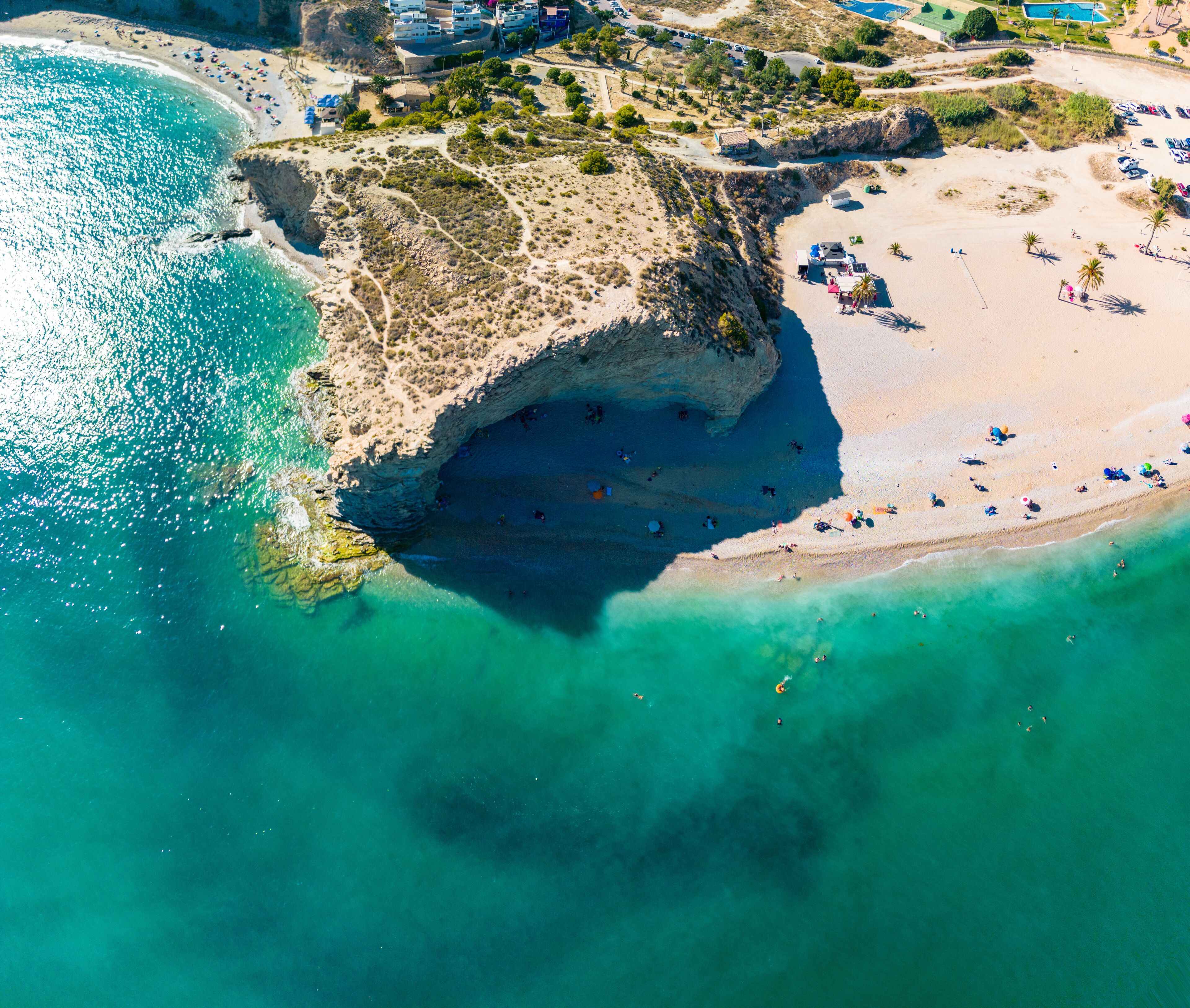 Vista aérea de una zona costera con playa, aguas turquesas cristalinas y un acantilado rocoso.